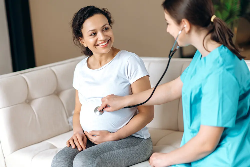 Happy pregnant woman and female doctor listening to baby's heartbeat sitting on a sofa